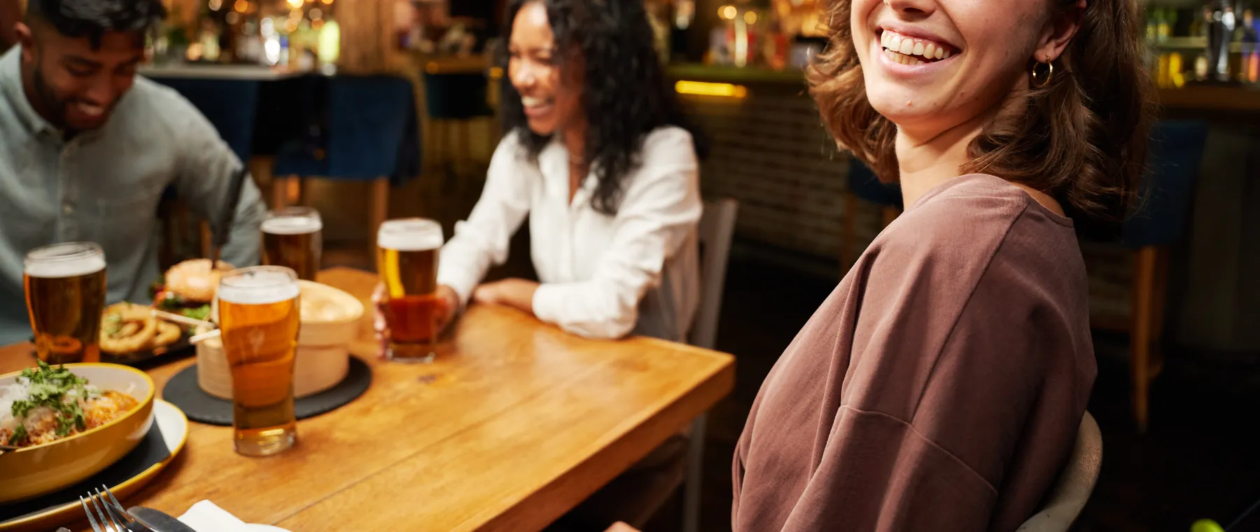 Gäste an einem Holztisch an der Bar eines Restaurants am Abend, vor ihnen Getränke und Essen