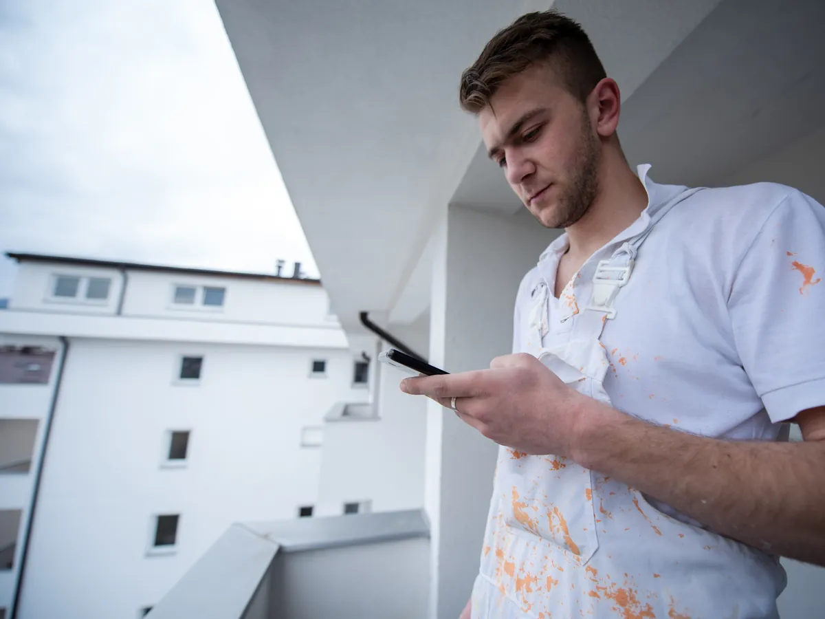 Maler in Arbeitskleidung schaut auf einem Balkon auf sein Smartphone