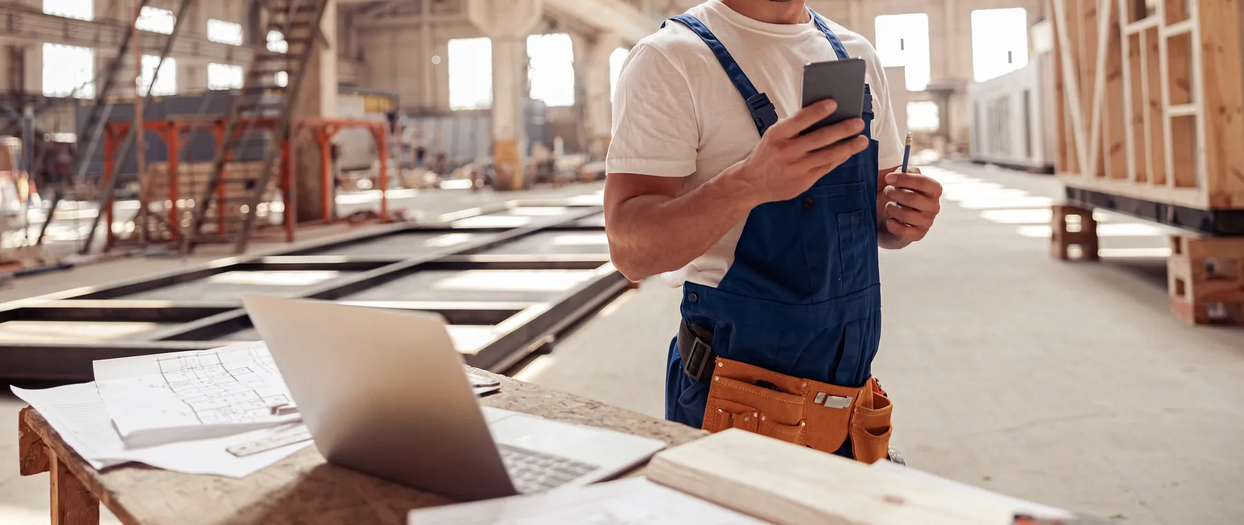 Handwerker in Latzhose und Helm bedient sein Smartphone in einer Zimmerei