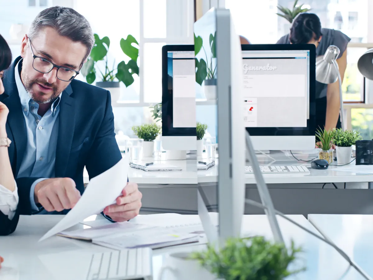 Steuerberater bespricht am Schreibtisch seiner Kanzlei Unterlagen, im Hintergrund die Arbeitsplätze des Teams